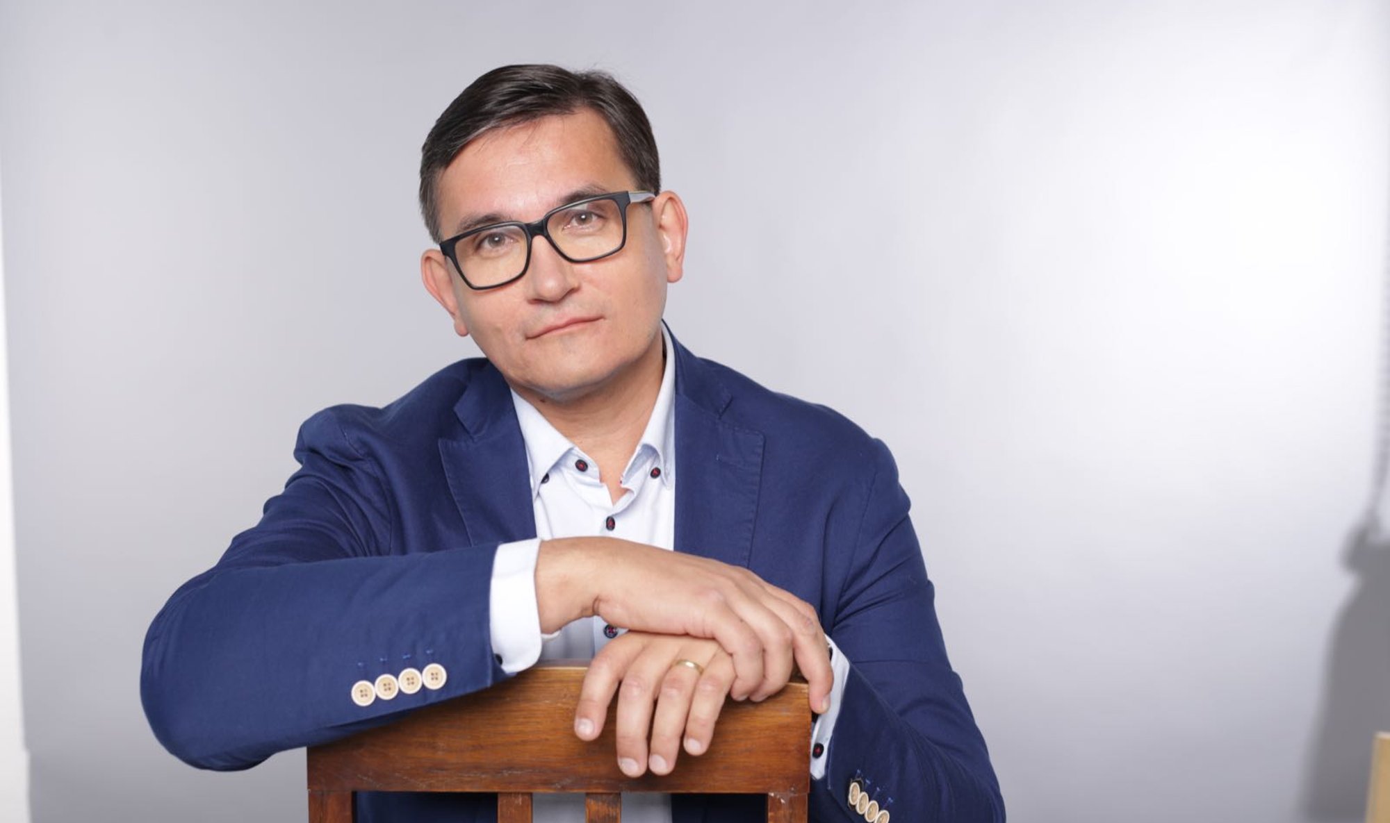 Person in a blue blazer and glasses leaning on a wooden chair.